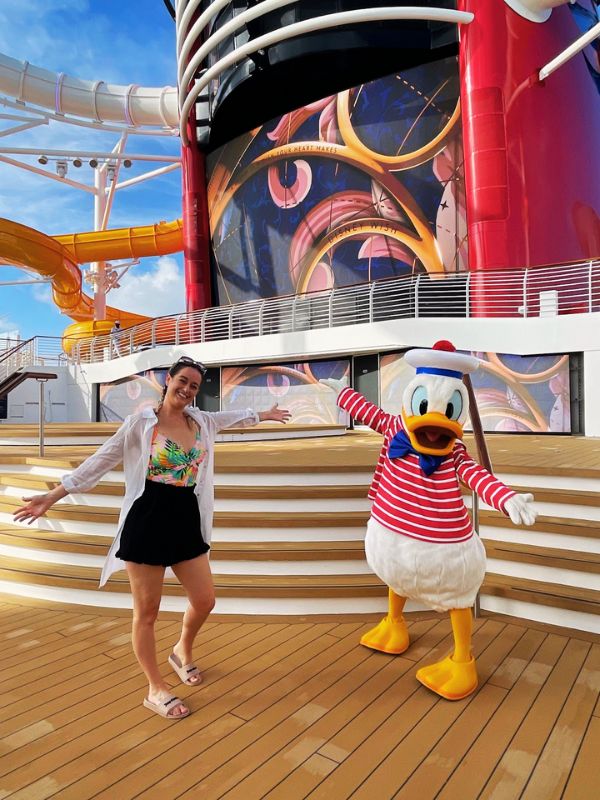 A joyful moment on a Disney cruise ship where Cruise Mummy is posing next to Donald Duck. They are standing on the ship's deck with a colorful backdrop featuring an artistic rendering of Mickey Mouse on the ship's funnel. She is dressed in a casual, tropical-themed outfit, smiling widely, matching Donald Duck's own animated pose in his classic sailor outfit. The sunny day and the ship's bright, playful design elements add to the cheerful, vacation atmosphere. This picture perfectly captures the family-friendly and fun-filled experience offered by Disney cruises.