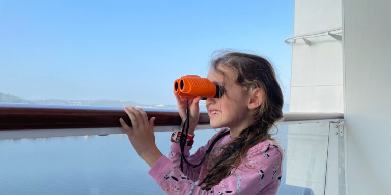 A young girl wearing a pink pajama top with black and white patterns is using orange binoculars to look out from a cruise ship balcony. She is smiling and appears excited, enjoying the scenic view of the calm water and distant coastline. The clear blue sky and peaceful surroundings enhance the moment of exploration.