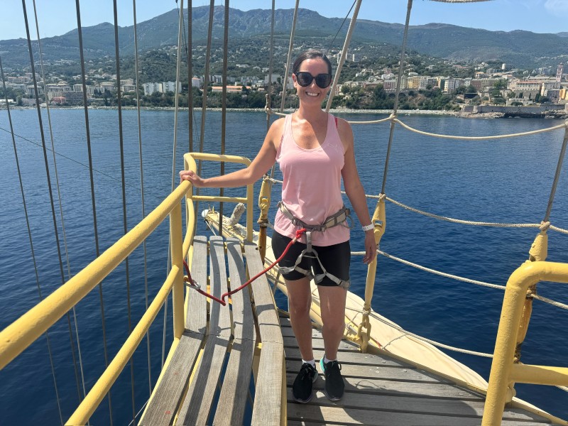 Cruise Mummy climbing the mast of a Star Clippers ship, standing on a wooden walkway surrounded by ropes and yellow railings. She is wearing a pink tank top, black shorts, and a safety harness while smiling confidently against a backdrop of calm blue water and a coastal city with hills. The scene highlights an adventurous and unique experience aboard a sailing vessel.