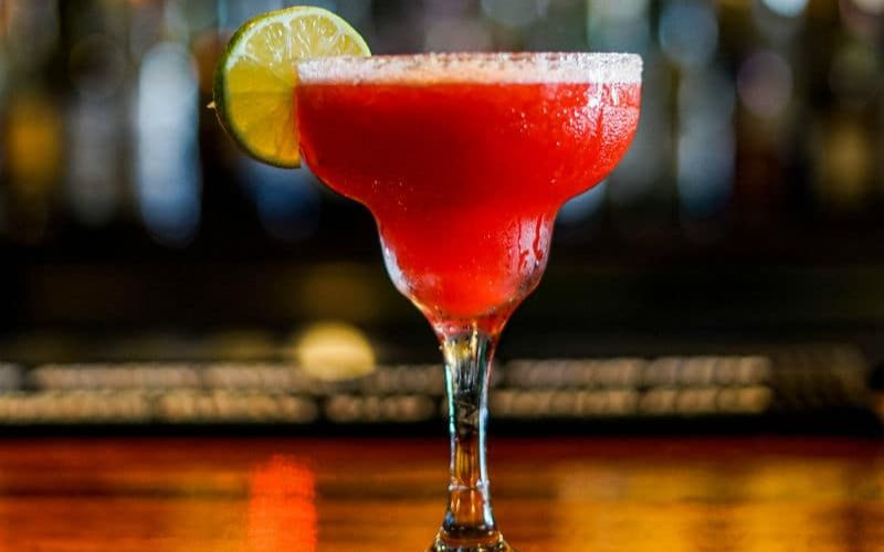 A vibrant red margarita served in a classic margarita glass with a salted rim. The drink is garnished with a slice of lime, adding a touch of citrusy freshness. The background is softly blurred, highlighting the colorful and refreshing cocktail, which sits on a polished wooden bar counter. The overall setting suggests a lively and inviting bar atmosphere, perfect for enjoying a delicious beverage.