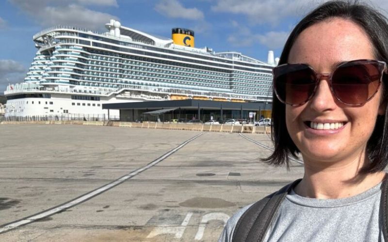 A woman with sunglasses is smiling in the foreground, with the large Costa cruise ship in the background, docked at a port under a partly cloudy sky with ample sunlight.
