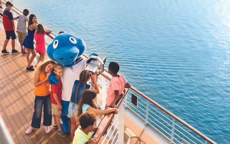 Group of children on a cruise ship deck enjoying the view of the ocean, accompanied by a person in a blue mascot costume. The children are interacting with the mascot and using binoculars to look out over the water, creating a lively and joyful atmosphere.