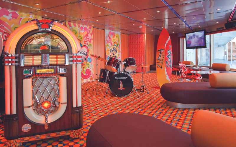 Retro-themed room on a cruise ship, featuring a classic jukebox, a drum set, and colorful decor with surfboards and vibrant patterns. The room has modern seating and a TV, creating a fun and nostalgic atmosphere perfect for music and entertainment activities.