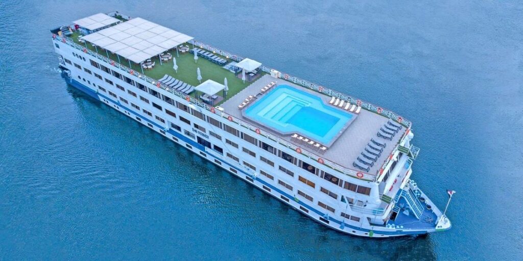An aerial view of the CroisiEurope river cruise ship, elegantly cruising on the water with a well-appointed sundeck featuring a large swimming pool. The ship's long, white structure with neat rows of windows is crowned with a spacious open-air lounge area, offering guests a luxurious and relaxing voyage.