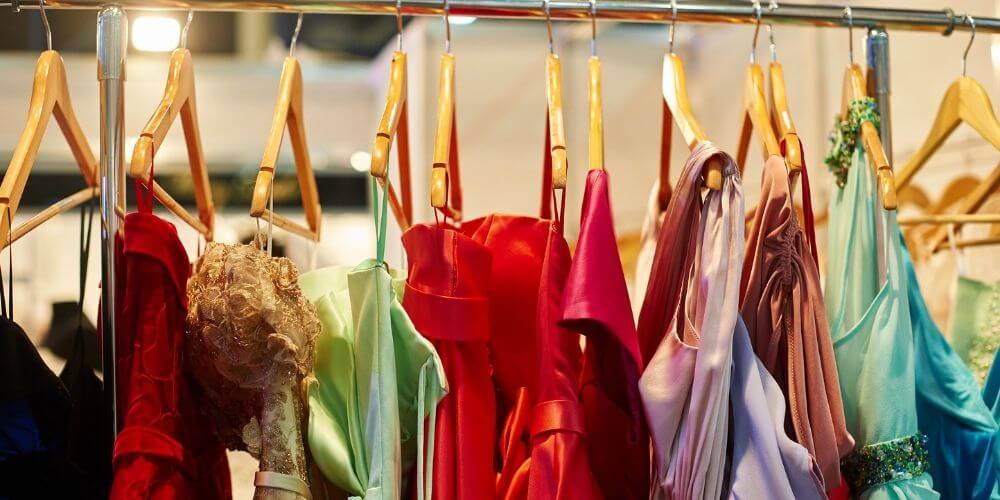 A rack of elegant evening dresses in various colors and styles, including red, green, pink, and blue, hanging on wooden hangers. The dresses are suitable for formal occasions on a cruise, showcasing a range of luxurious fabrics and designs.
