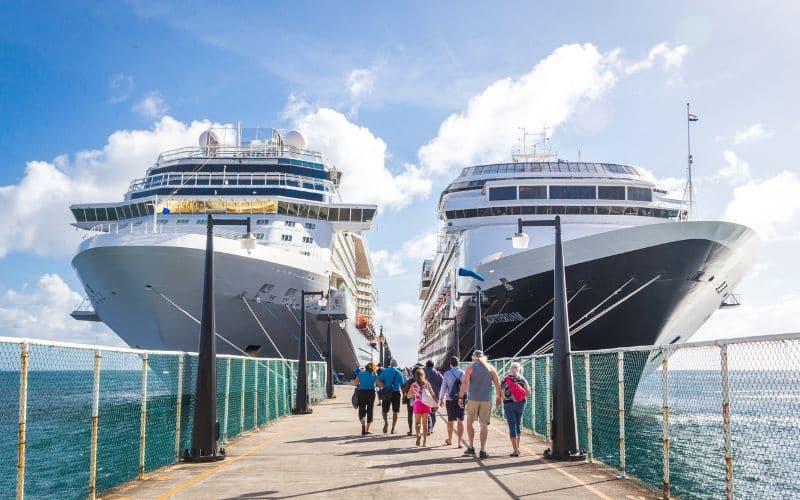 Two large cruise ships docked side by side at a port, with people walking along the pier under a bright, sunny sky.