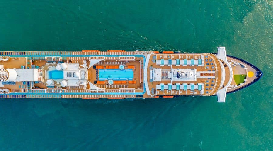 Aerial view of a large cruise ship sailing on vibrant green ocean waters. The top deck is bustling with amenities including two large swimming pools, rows of lounge chairs, and a variety of colorful recreational areas, showcasing the ship’s leisure and entertainment facilities.