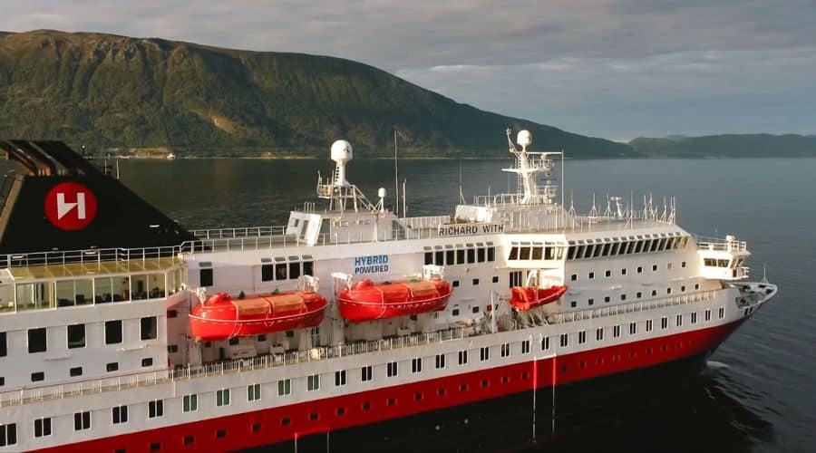 A hybrid-powered cruise ship named 'Richard With' sails close to a lush, mountainous coastline. The ship features a distinctive black funnel with a red logo, and its hull is painted in white and red. Lifeboats are mounted on the side, ready for emergency use, reflecting the ship's modern safety measures and sustainable design.