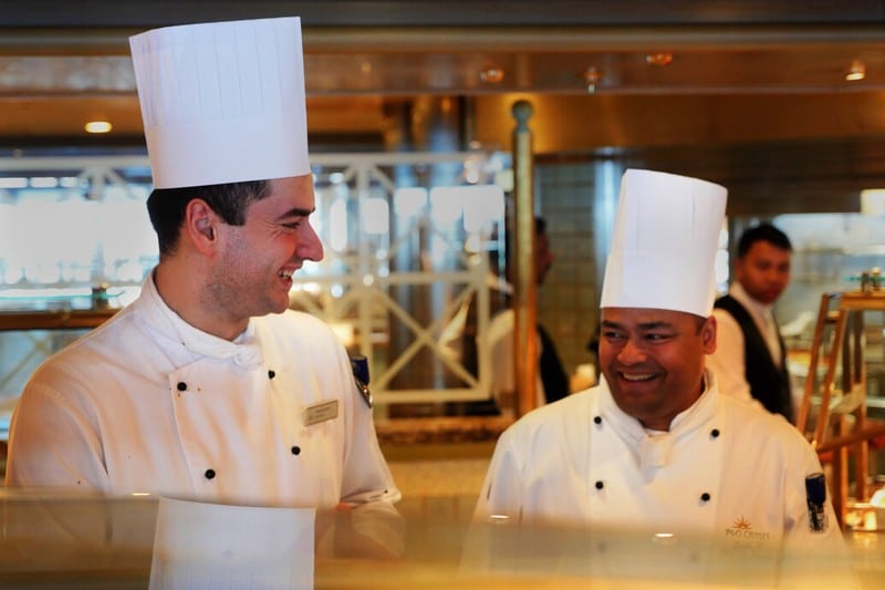 Two P&O Cruises' chefs, wearing traditional white uniforms and tall chef hats, are smiling and conversing in the ship's kitchen. They are surrounded by the gleaming stainless steel of the kitchen equipment and the warm lighting of the galley. The chefs' friendly interaction reflects a positive and professional working environment.