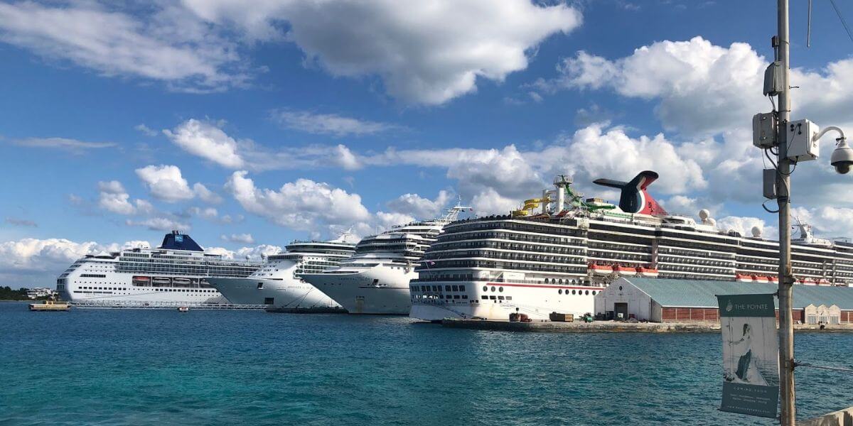 Several large cruise ships docked at a port under a bright, partly cloudy sky. The clear blue water and the bustling harbor highlight the busy and vibrant cruise industry.