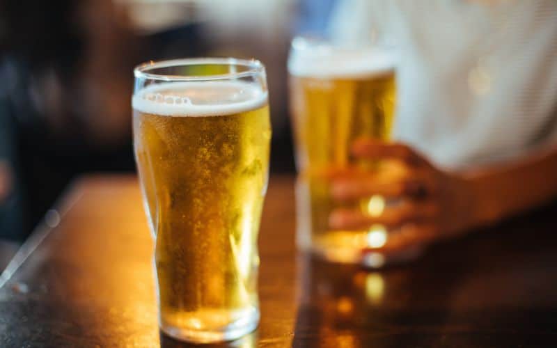 Two pint glasses of beer with a perfect head of foam, one in the foreground and one held by a person in the background, on a wooden surface with a warm, inviting ambiance typical of a cozy pub or social gathering area on a cruise.