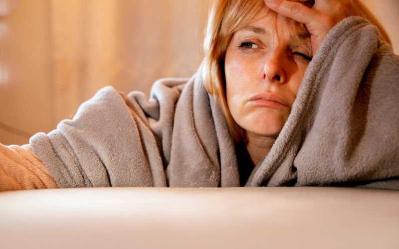 A woman wearing a plush grey robe looks drowsy and fatigued, resting her head on her hand. Her tired expression and the soft, warm lighting suggest she might be feeling unwell or exhausted.