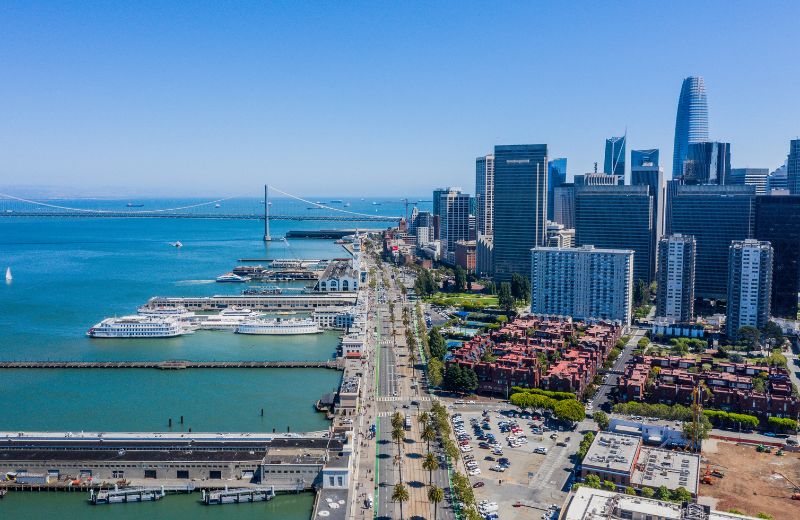 The vibrant Embarcadero waterfront area of San Francisco, showcasing a sweeping view of the cityscape along with the iconic Bay Bridge stretching into the horizon. The Embarcadero is bustling with activity, visible from the cars on the road and people along the sidewalks, adjacent to the historic Ferry Building and various cruise ships docked at the piers. The backdrop features a mix of modern skyscrapers and classic San Francisco architecture, highlighting the dynamic urban landscape. This setting is a popular spot for both tourists and locals, offering scenic views, dining, and shopping opportunities, and acts as a critical transport hub in the city.