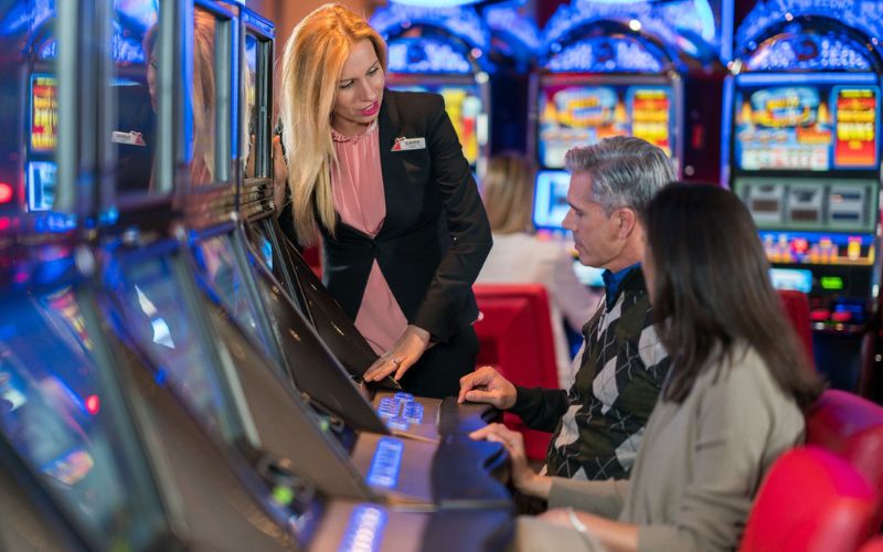 A cruise ship casino staff member is assisting a couple seated at a row of slot machines. The staff member, dressed in a formal blazer, is attentively explaining something to the guests, who are focused on the machines. The casino is brightly lit with colorful lights and displays from the various slot machines, creating an exciting and engaging atmosphere.