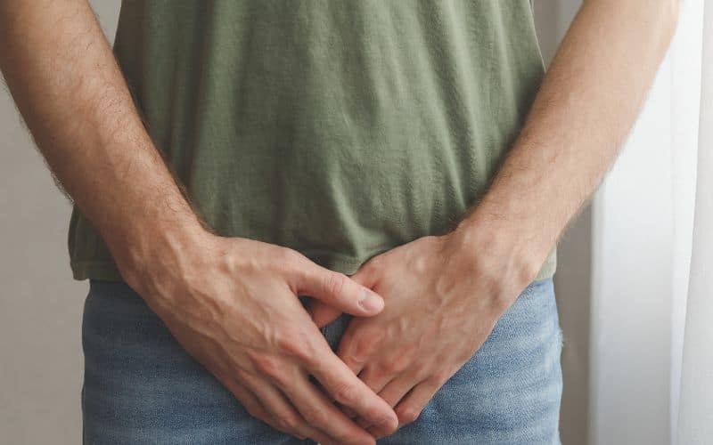 A person in a green t-shirt and jeans covering their groin area with both hands, possibly indicating discomfort or protection. The image is cropped to show only the torso and hands.