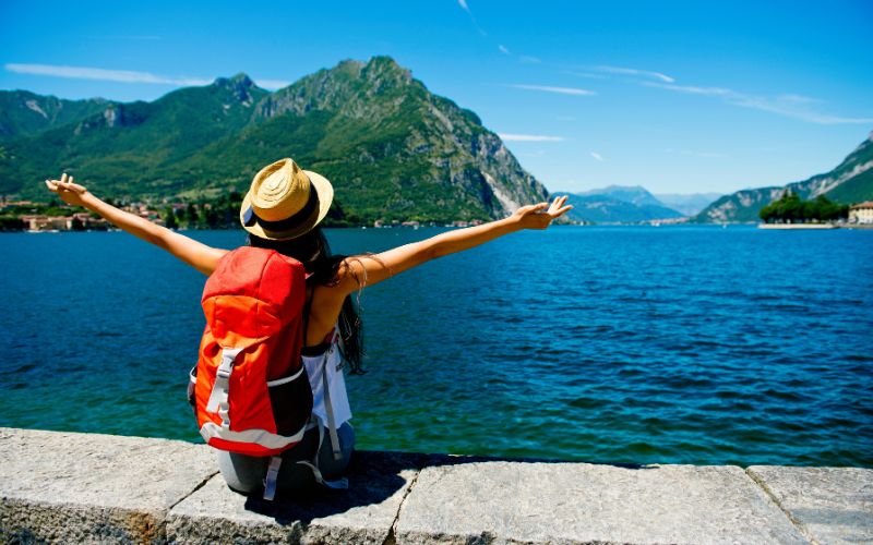 A female traveler wearing a hat and backpack sits on a stone ledge with her arms outstretched, facing a serene lake surrounded by mountains. The bright, clear day highlights the natural beauty and sense of adventure.