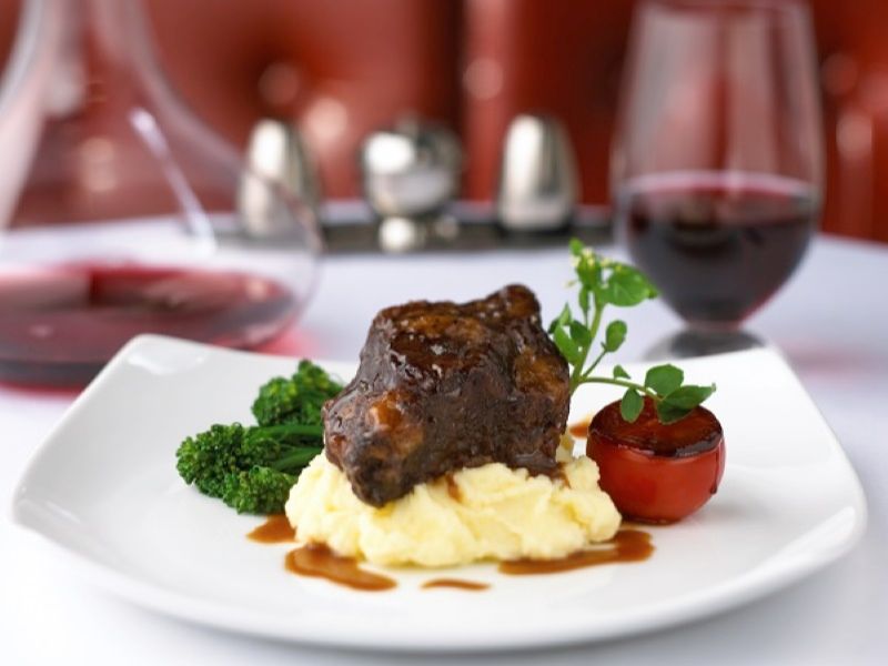 A succulent braised short rib dish from Chops Grille on Anthem of the Seas, elegantly presented on a white plate with mashed potatoes, broccolini, and a glazed tomato, accompanied by a glass of red wine in the background.