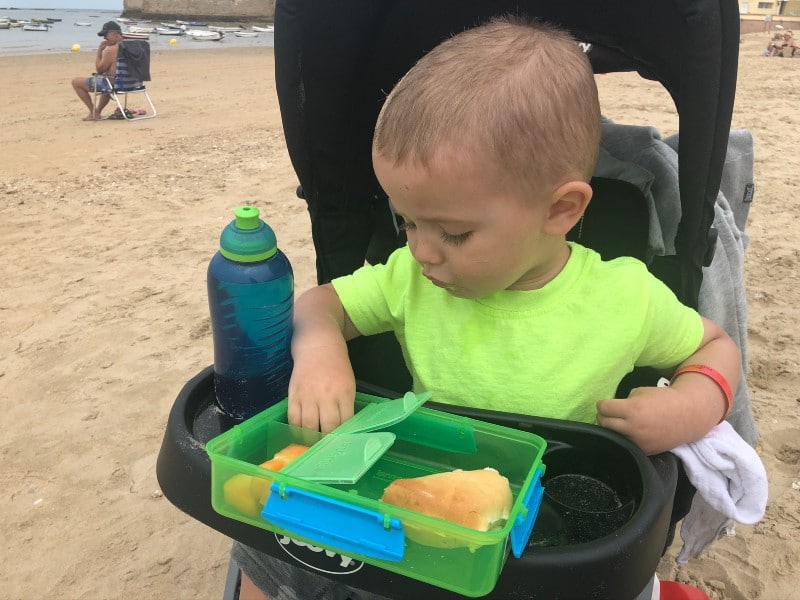 A toddler in a neon green shirt seated in a stroller on the beach, focused on a green lunchbox with a sandwich and snacks, with a blue water bottle beside them, and beachgoers in the background enjoying a cloudy day.