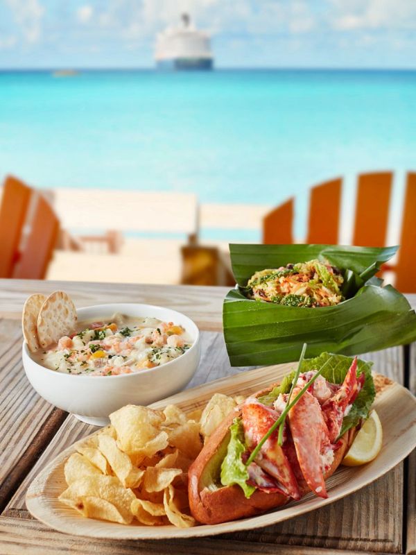 A delicious seafood meal is presented in the foreground with a lobster roll, chips, and a creamy shrimp salad, served on a wooden platter. The tropical ambiance is enhanced by the blurred cruise ship in the background, suggesting a luxurious dining experience by the vibrant turquoise waters.