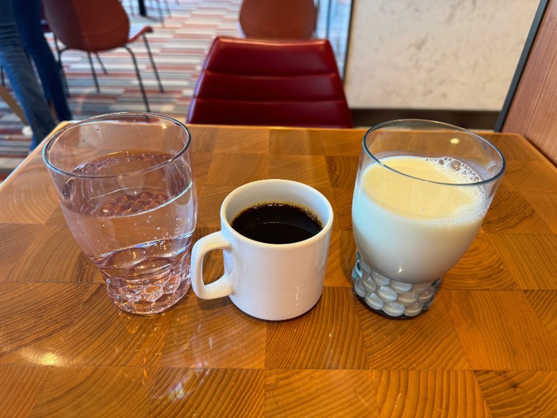 A table setting featuring a glass of water, a cup of black coffee, and a glass of milk, all offered as free drinks on a wooden table in a café. The background includes red and brown chairs, emphasizing the casual and welcoming atmosphere of the dining area.