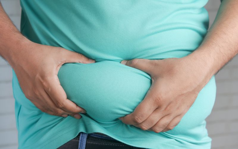 This image features a close-up of a man holding his stomach, which is slightly bulging under a teal t-shirt. His hands are gently wrapped around the sides of his belly, emphasizing the fullness. The setting appears to be casual with a blurred brick wall background, possibly focusing on themes of weight gain or body image.