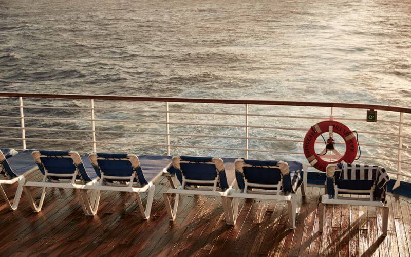 The image shows a row of empty lounge chairs lined up on the lido deck of the Grand Princess cruise ship, facing the open sea. The wooden deck gleams with a warm, sunlit hue, and a life preserver with "Grand Princess" is mounted on the railing. The tranquil ocean in the background complements the relaxed, inviting setup of the deck chairs.
