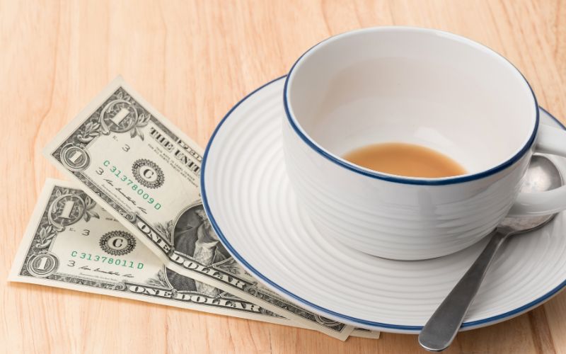An empty teacup with a small amount of tea remaining, placed on a saucer with a spoon, and two one-dollar bills underneath the saucer, symbolizing a gratuity left for service.