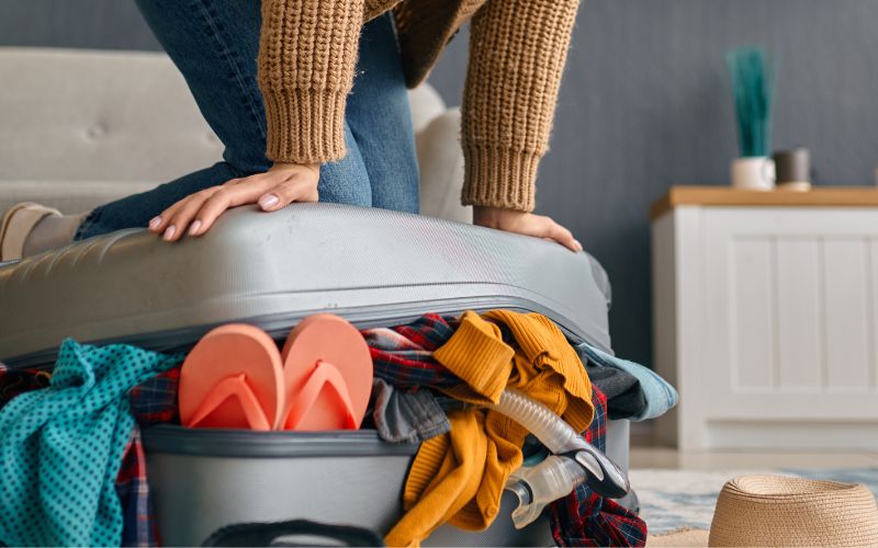 A woman struggles to fit her shoes into an overloaded suitcase, showcasing a chaotic packing scene.