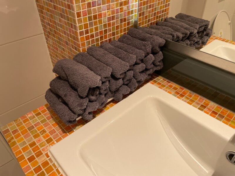 A bathroom sink area with an orange and yellow mosaic tile backsplash. A stack of neatly folded dark gray towels is placed on the counter next to the sink and mirror.