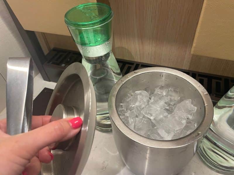 This image shows a metal ice bucket filled with ice cubes next to a clear glass carafe with a green lid, placed on a countertop. A hand with pink nail polish is holding the lid of the ice bucket.