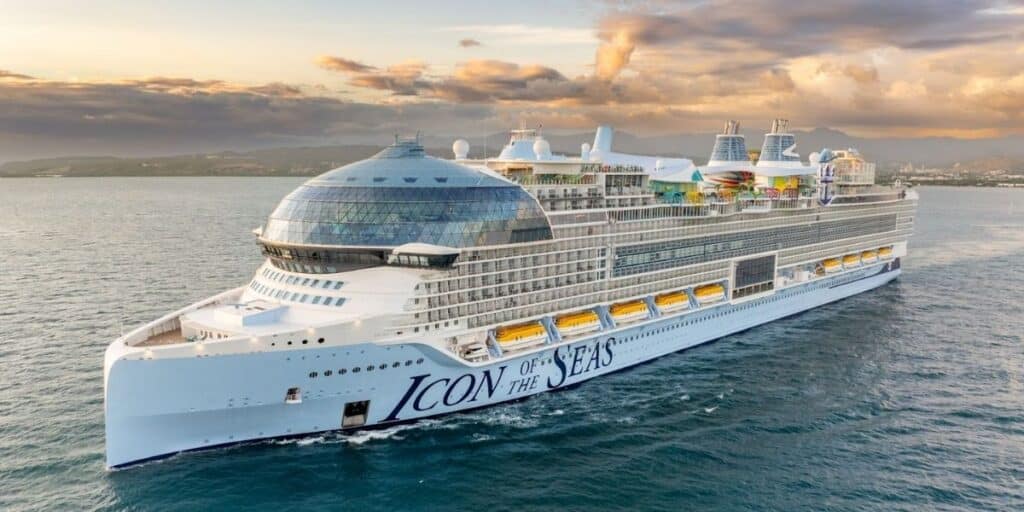 The 'Icon of the Seas' cruise ship by Royal Caribbean sails on calm seas during a vibrant sunset, with a coastal landscape in the background. This massive white ship features an array of amenities visible on the top decks, including colorful water slides and outdoor pools.