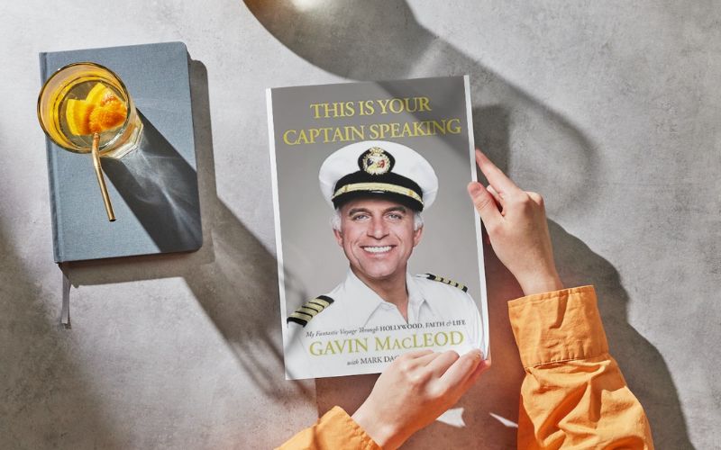 A person's hands holding a book titled 'This is Your Captain Speaking' by Gavin MacLeod, with a portrait of the author in a captain's uniform on the cover. Next to the book is a blue journal and a glass of water with a slice of orange, all laid out on a textured surface with sunlight casting a soft shadow.