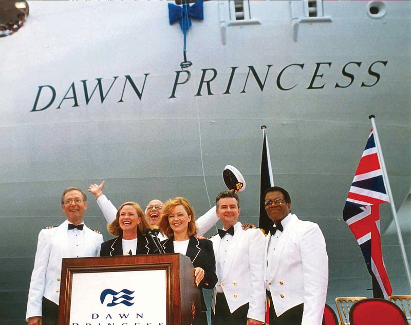 Lauren Tewes and The Love Boat cast in white naval uniforms in front of the 'Dawn Princess' cruise ship. They are gathered around a podium, waving and smiling, with maritime flags and the ship's name prominently displayed in the background.