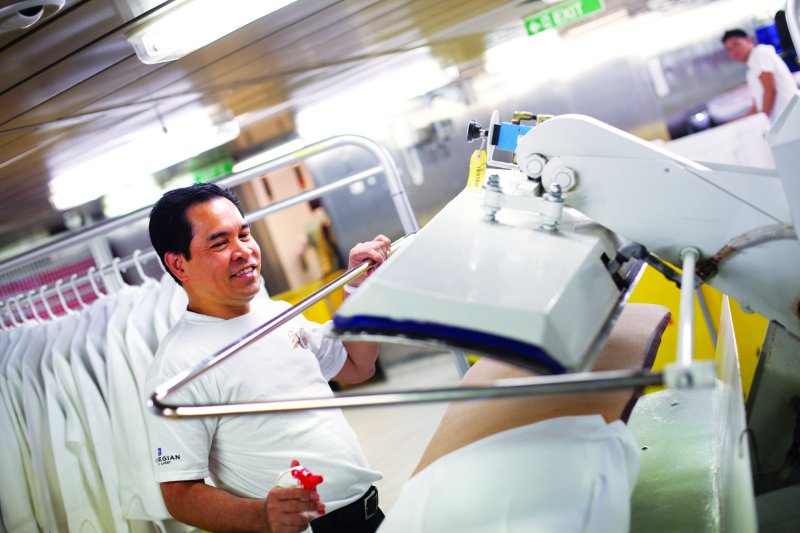 A cheerful crew member operates a large industrial ironing machine, pressing white linens in the laundry room of a cruise ship, with a focus on impeccable onboard service.