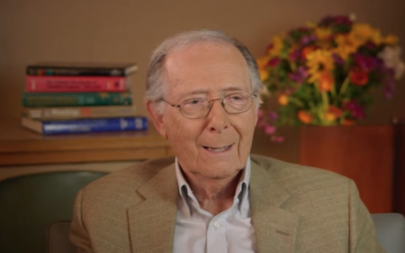 Bermie Kopell smiling warmly, is seated in an indoor setting for an interview. He's wearing a beige jacket over a white shirt. In the background, there's a shelf with a stack of books and a vase of vibrant yellow and purple flowers.