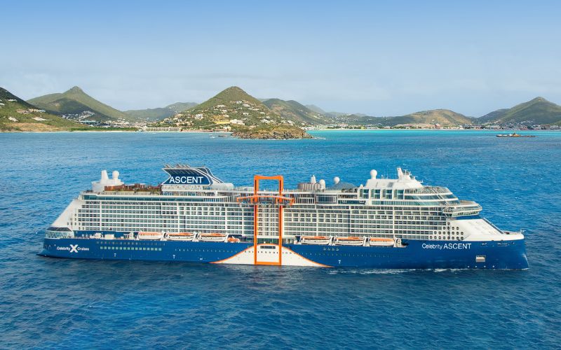 The 'Celebrity Ascent' cruise ship, captured in clear weather, cruising near a coastal landscape with lush green hills. Its distinctive blue hull and orange accents highlight the ship's elegant design as it traverses the turquoise waters.