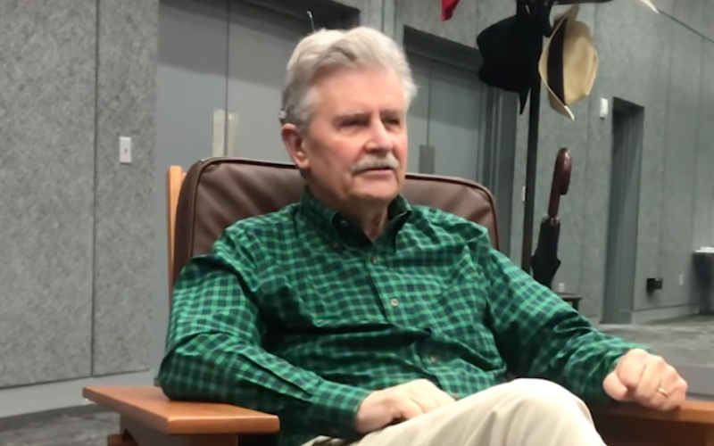 Fred Grandy, with distinguished gray hair and a mustache sits comfortably in a brown leather office chair. He's wearing a green plaid shirt and khaki pants, giving an attentive look.