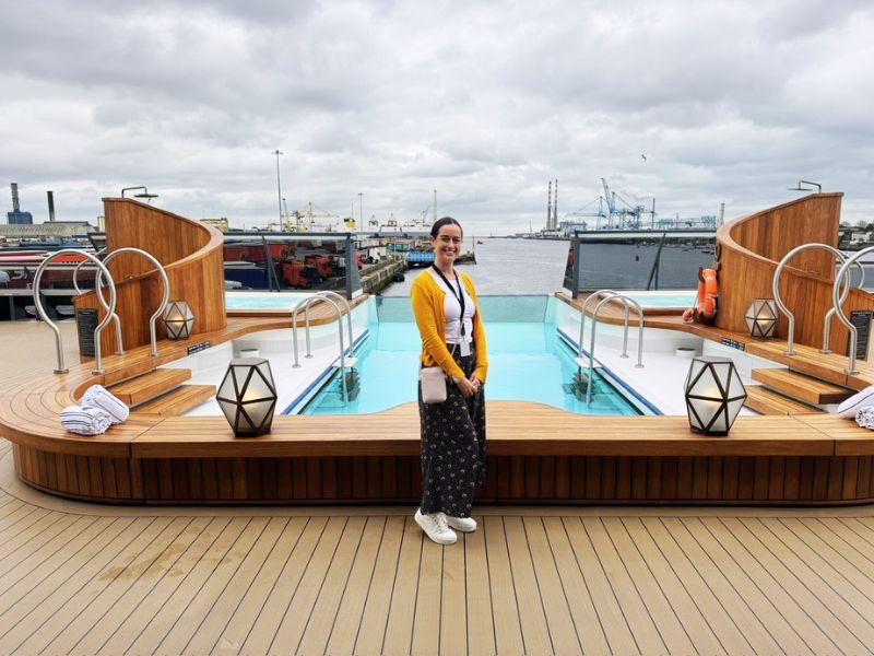 Cruise Mummy stands on the deck of the Seabourn Venture, smiling in front of the ship's inviting infinity pool and two spacious hot tubs. The wooden deck curves around the water features, which are accentuated by elegant metal railings and sophisticated deck furniture, including plush towels and a geometric lantern. The port setting in the background, with its industrial cranes and docks, contrasts with the luxurious and serene ambiance of the ship’s outdoor relaxation area.