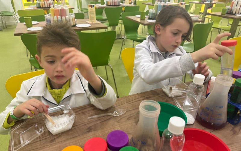 Two young children dressed in white lab coats are engaged in a fun science activity, experimenting with various bottles and colored solutions at a bright and colorful table, fostering an early interest in science and experimentation.