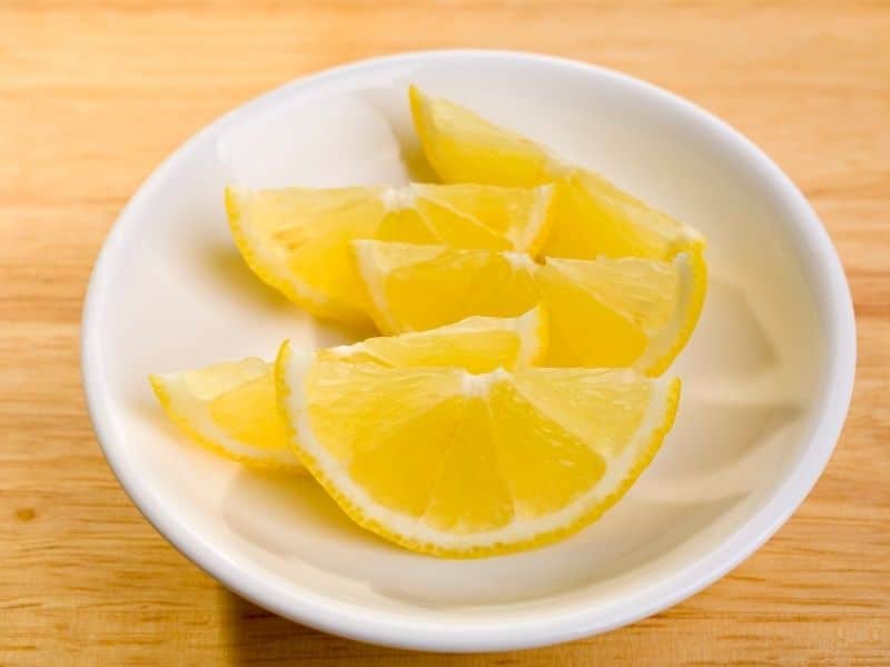 This image shows a small white dish containing several fresh lemon slices arranged neatly. The bright yellow lemons contrast with the clean white plate, set on a light wooden surface. The presentation highlights the vibrant color and juiciness of the lemons.