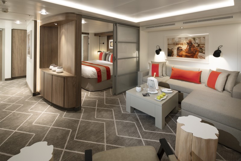 Elegant Celebrity Suite on a Celebrity cruise ship, featuring a sophisticated living area with a gray sofa accented with red pillows, a contemporary coffee table, stylish artwork, and a glimpse into the cozy bedroom area.