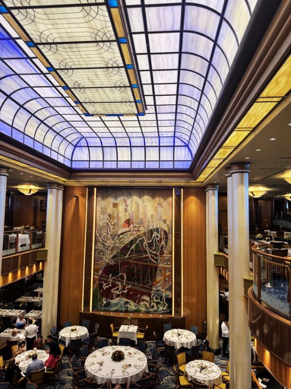The main dining room on the Cunard Queen Mary cruise ship features an elegant, two-story space with a grand stained-glass ceiling, large mural, and marble columns. The dining area is set with round tables adorned with white tablecloths and floral centerpieces, creating a refined and luxurious dining atmosphere.