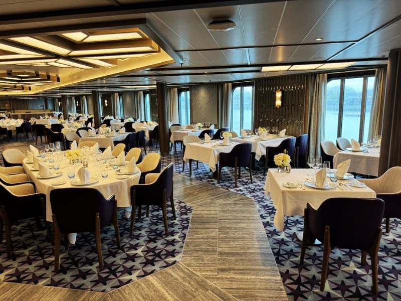 The main dining room on the Seabourn Venture exudes elegance and sophistication with its spacious layout. The room is arranged with neatly set round and rectangular tables, adorned with white tablecloths, fine china, and yellow floral centerpieces. The stylish decor includes plush blue and cream chairs, patterned carpet flooring, and modern geometric lighting, creating an inviting atmosphere for dining with views of the ocean through large windows.
