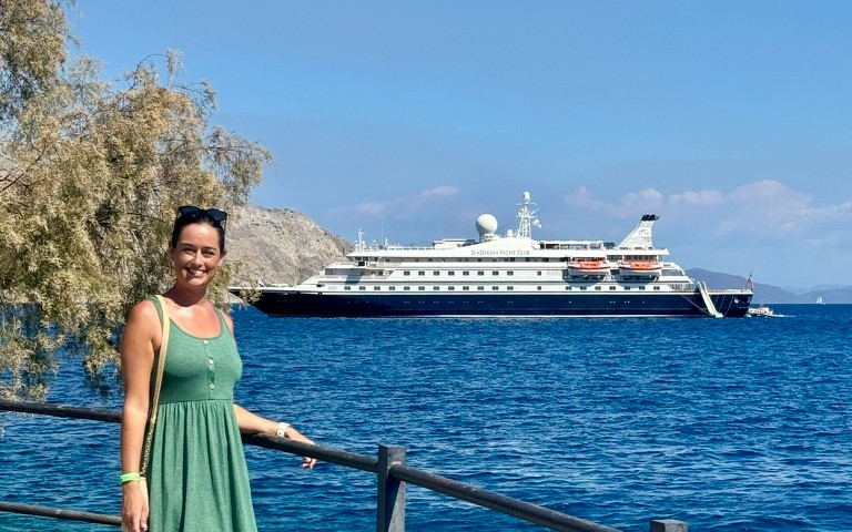 Cruise Mummy standing near a railing with a backdrop of a SeaDream Yacht Club vessel anchored in a tranquil blue sea. She is smiling, dressed in a green sleeveless dress, enjoying the sunny weather, with the yacht and distant hills completing the scenic, relaxed atmosphere. The composition highlights the luxurious and intimate setting of a yacht cruise.