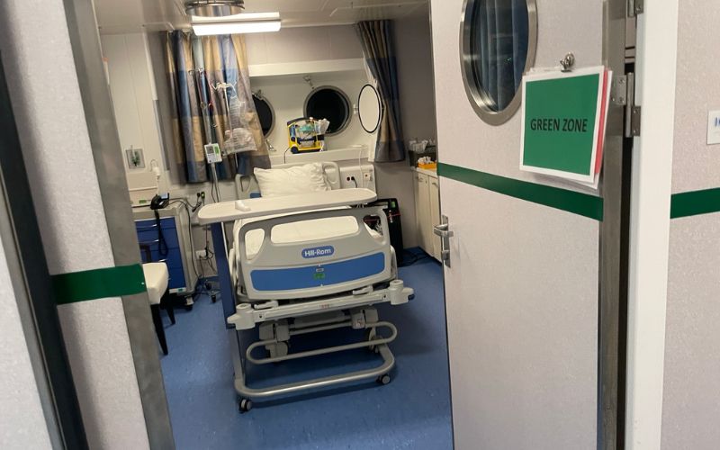 The image shows an inside view of a hospital room, viewed from the doorway. It features a modern Hill-Rom hospital bed centered in the room, with medical curtains partially drawn around it. To the right of the door, there's a sign reading "GREEN ZONE". The environment appears sterile and well-equipped for patient care.
