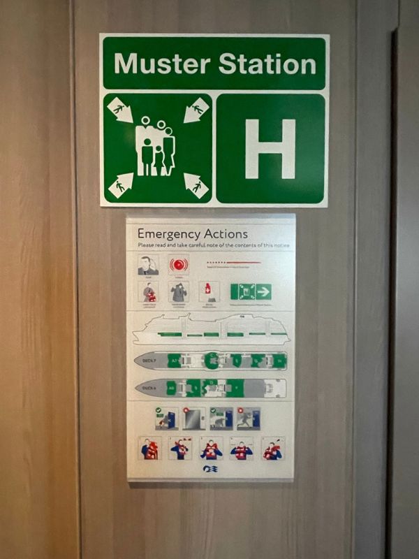 The image shows a muster station sign on a cruise ship, labeled with the letter "H" and featuring a green background with safety icons. Below it is an emergency actions guide, which includes instructions and diagrams for passengers on how to proceed in case of an emergency, as well as maps of the ship's decks and muster points.