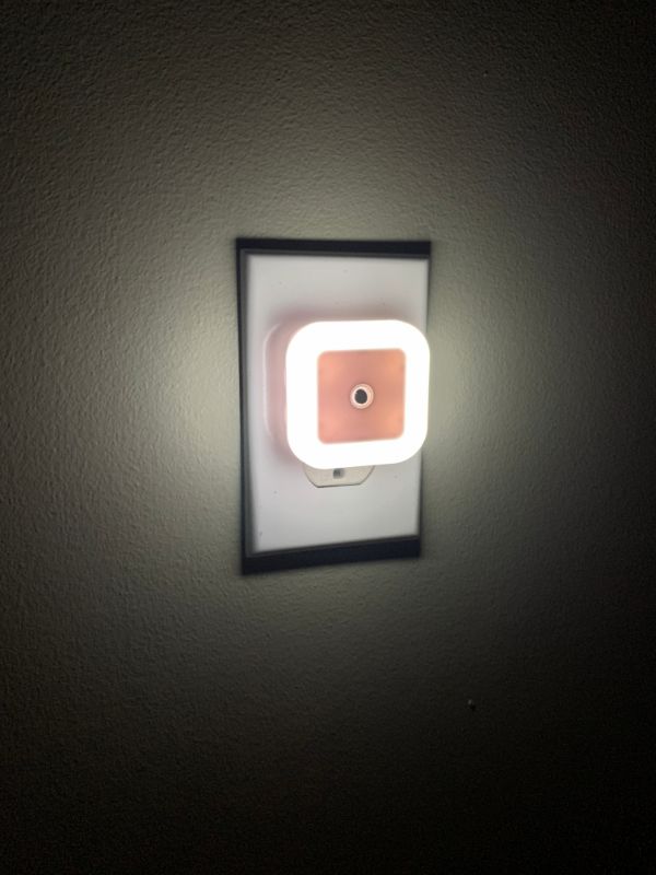 A small, square-shaped nightlight is plugged into an electrical outlet on a wall, emitting a soft white glow. The light provides gentle illumination in the darkened room, creating a cozy and safe atmosphere.
