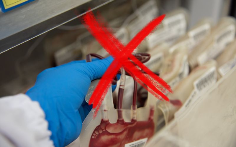 This image displays a hand in a blue glove pointing to a vial of blood in a laboratory setting, with a prominent red cross mark superimposed on the image, suggesting rejection or ineligibility. The shelves in the background hold multiple other samples, highlighting a medical or research environment where blood testing or collection is being conducted. The red cross might indicate a specific sample that is unsuitable for use or testing.