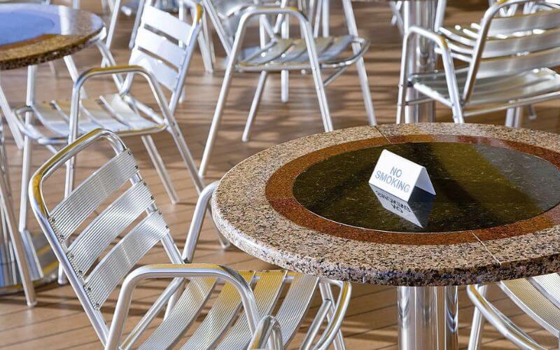 A 'No Smoking' sign placed on a wet circular table with a mosaic trim, surrounded by metal chairs on the wooden deck of a cruise ship, conveying a clear non-smoking policy in the outdoor seating area.