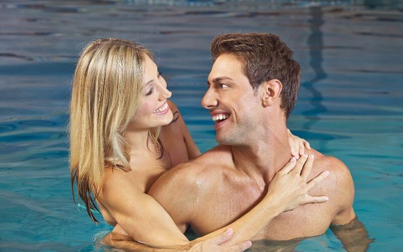 A smiling couple embracing in a swimming pool, looking at each other with joy. The woman has blonde hair and the man has dark hair, both appearing happy and relaxed in the water.